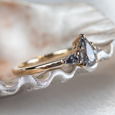 A gold three-stone ring with a fancy grey pear-shaped diamond center and two smaller grey pear-shaped side diamonds on a metallic, textured surface.