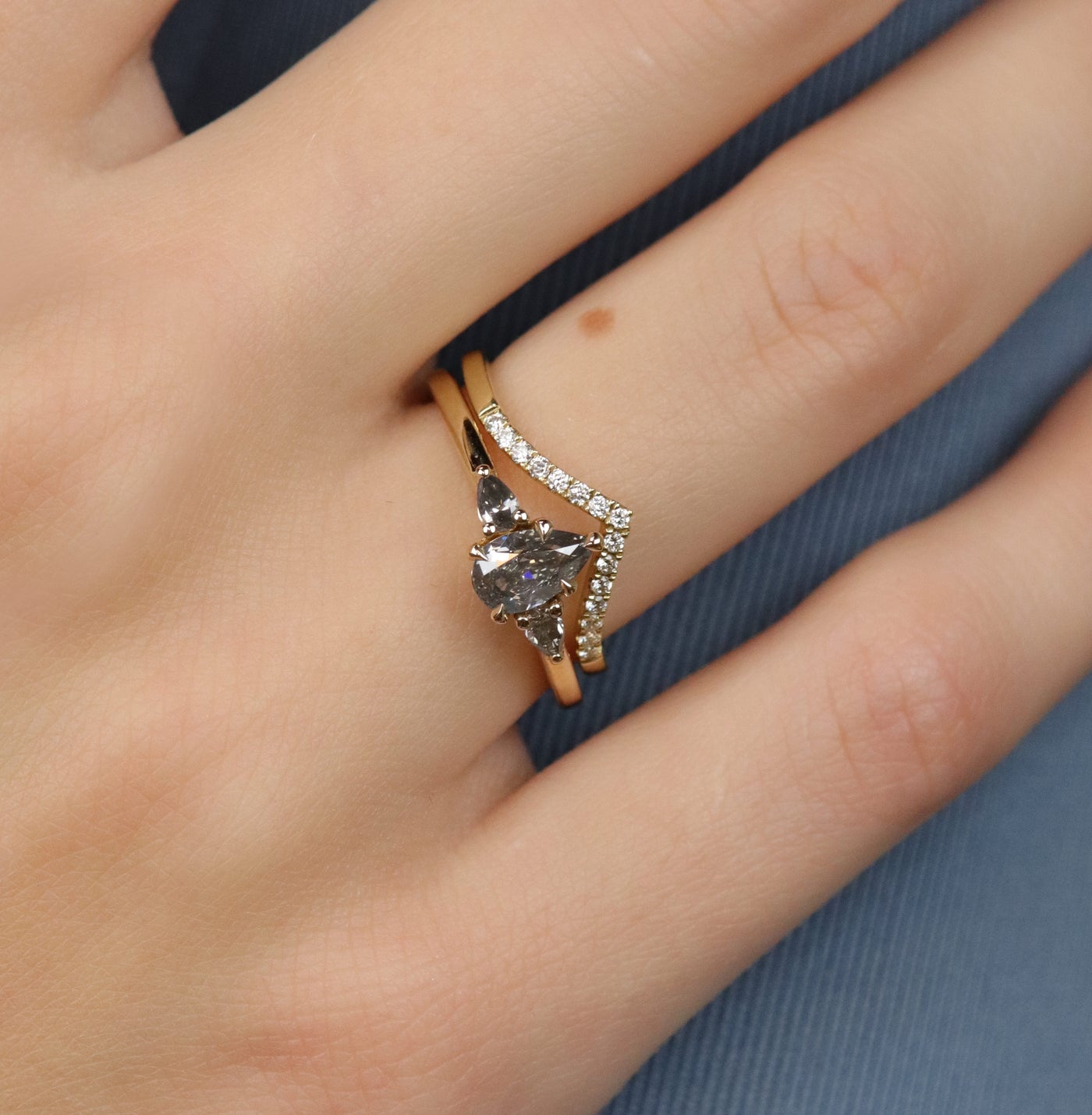 A gold three-stone ring with a fancy grey pear-shaped diamond center and two smaller grey pear-shaped side diamonds, stacked with a 'V' shaped diamond band, worn on a hand.