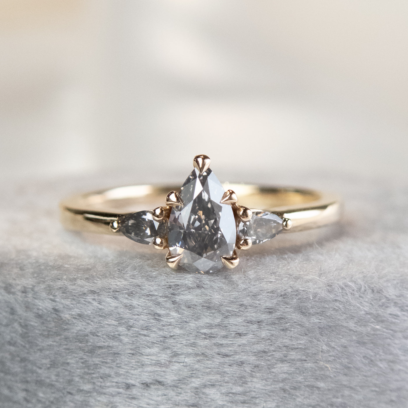 A gold ring with a fancy grey pear-shaped diamond center and two smaller grey pear-shaped side diamonds on a soft, textured surface.