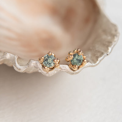 A pair of light teal green sapphire baleal stud earrings in 14KT yellow gold, displayed on a white surface.
