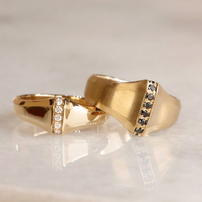 Two gold rings, one shiny with a central line of white diamonds and the other with a matte finish featuring a central line of black diamonds shown resting together on a white marble surface.