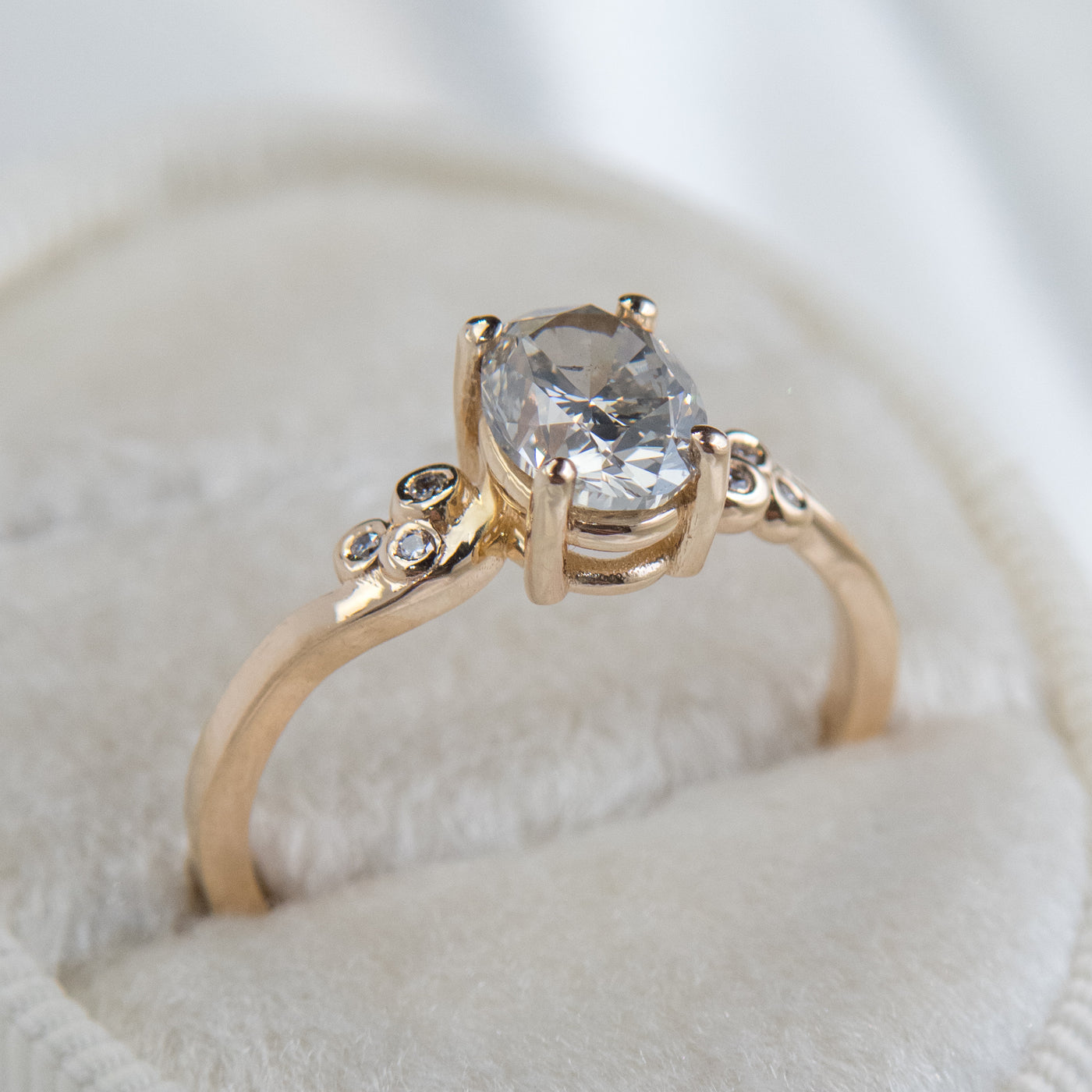 A gold ring with a fancy grey oval diamond at center, flanked by three small round diamonds on each side, lies on a soft white surface.