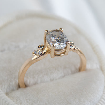A gold ring with a fancy grey oval diamond at center, flanked by three small round diamonds on each side, lies on a soft white surface.