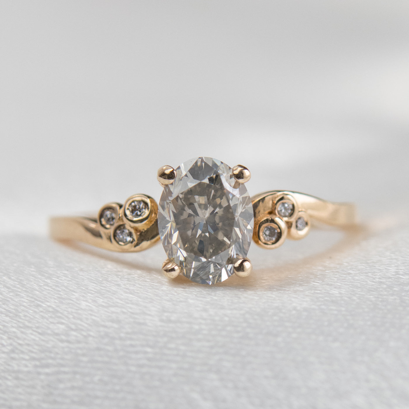 A gold ring with a fancy grey oval diamond at center, flanked by three small round diamonds on each side, lies on a soft white surface.