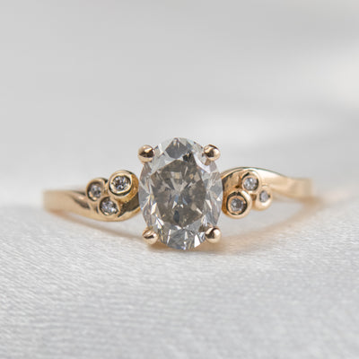 A gold ring with a fancy grey oval diamond at center, flanked by three small round diamonds on each side, lies on a soft white surface.