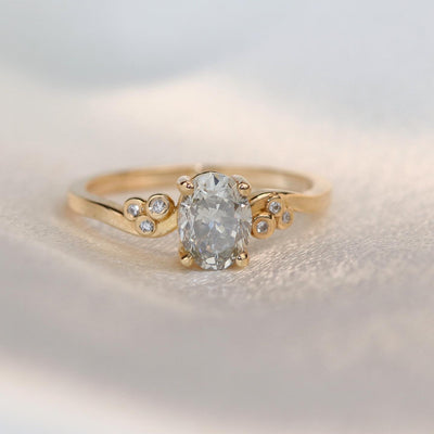 A gold ring with a fancy grey oval diamond at center, flanked by three small round diamonds on each side, lies on a soft white surface.