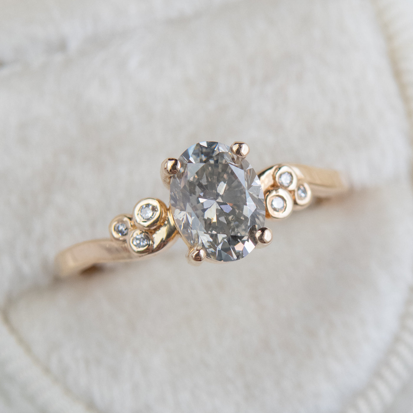A gold ring with a fancy grey oval diamond at center, flanked by three small round diamonds on each side, lies on a soft white surface.