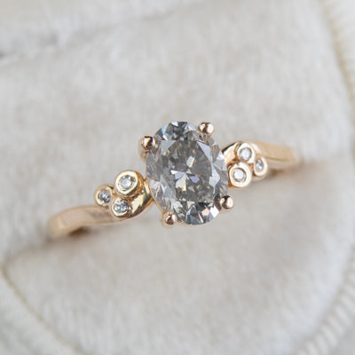 A gold ring with a fancy grey oval diamond at center, flanked by three small round diamonds on each side, lies on a soft white surface.