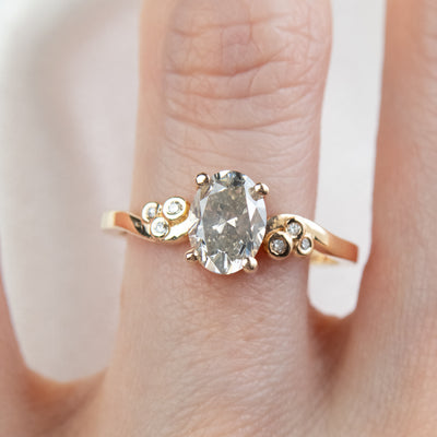 A gold ring with a fancy grey oval diamond at center, flanked by three small round diamonds on each side, worn on a finger. 