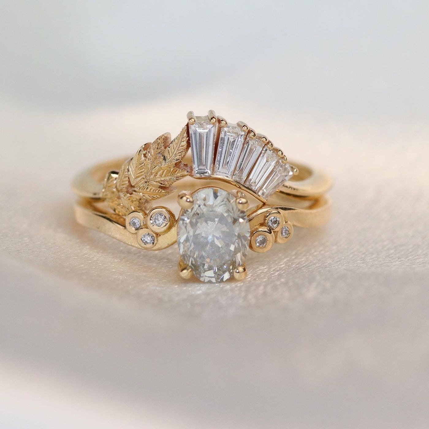 Two gold rings, one featuring a central fancy grey oval diamond, the other with a vertical row of baguette diamonds and leaf detail, conveying a vintage and luxurious feel. 
