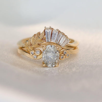 Two gold rings, one featuring a central fancy grey oval diamond, the other with a vertical row of baguette diamonds and leaf detail, conveying a vintage and luxurious feel. 