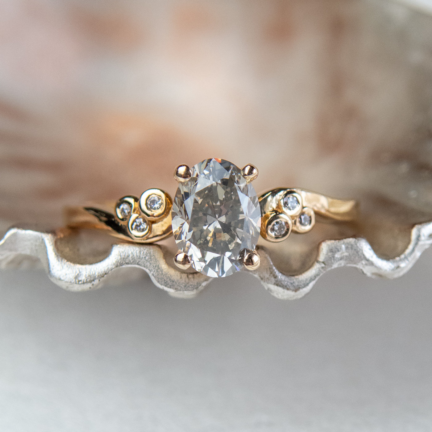 A gold ring with a fancy grey oval diamond at center, flanked by three small round diamonds on each side, lies on a metallic, shell-like surface. 