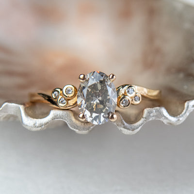 A gold ring with a fancy grey oval diamond at center, flanked by three small round diamonds on each side, lies on a metallic, shell-like surface. 