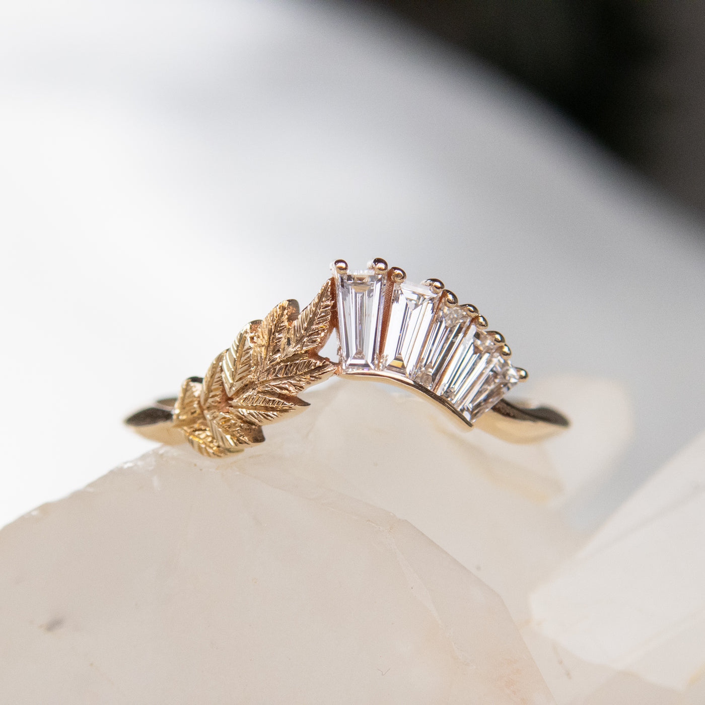 A slightly curved gold ring featuring a hand engraved leaf design and a row of sparkling, baguette cut lab grown diamonds resting on a crystal surface.