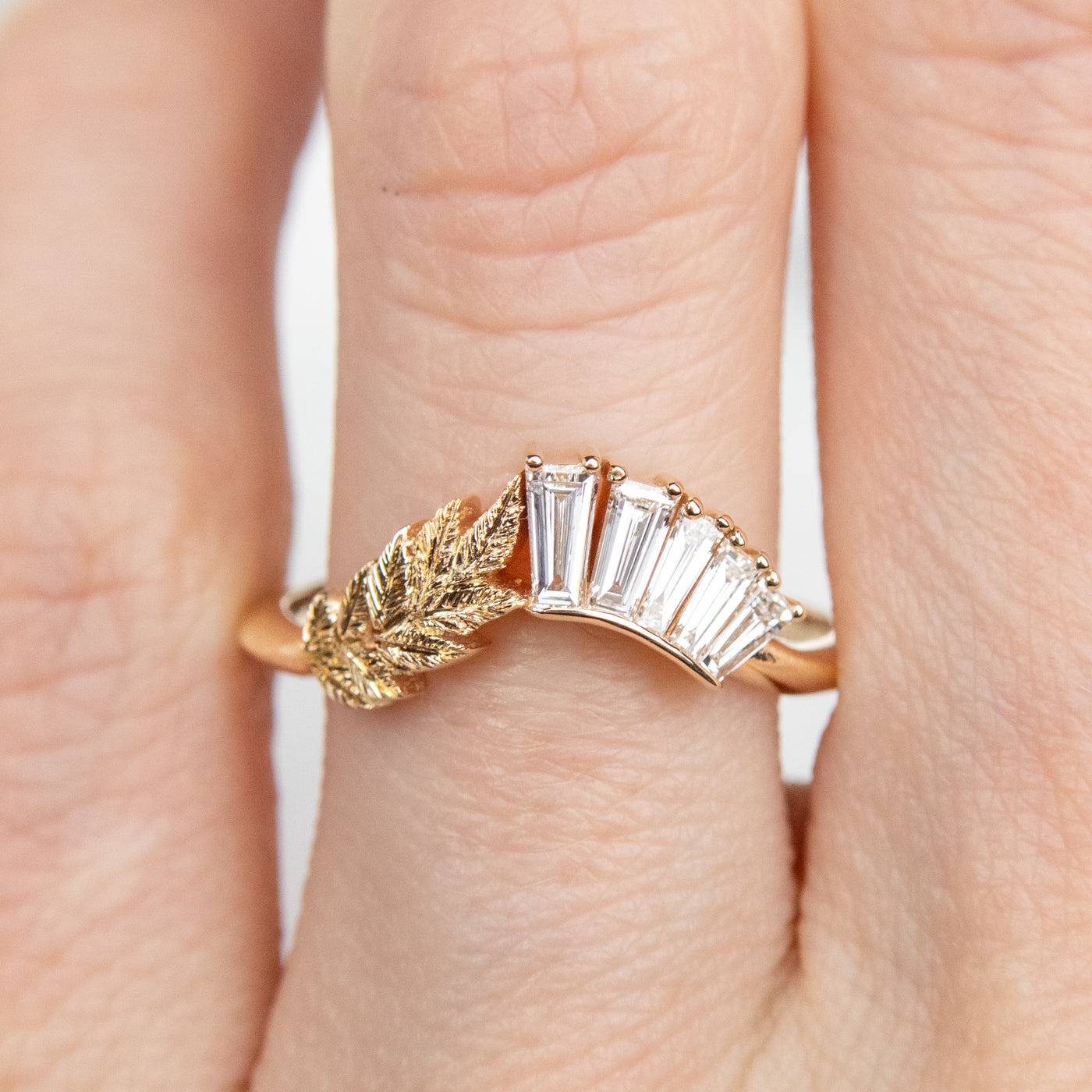 A slightly curved gold ring featuring a hand engraved leaf design and a row of sparkling, baguette cut lab grown diamonds shown worn on a finger.