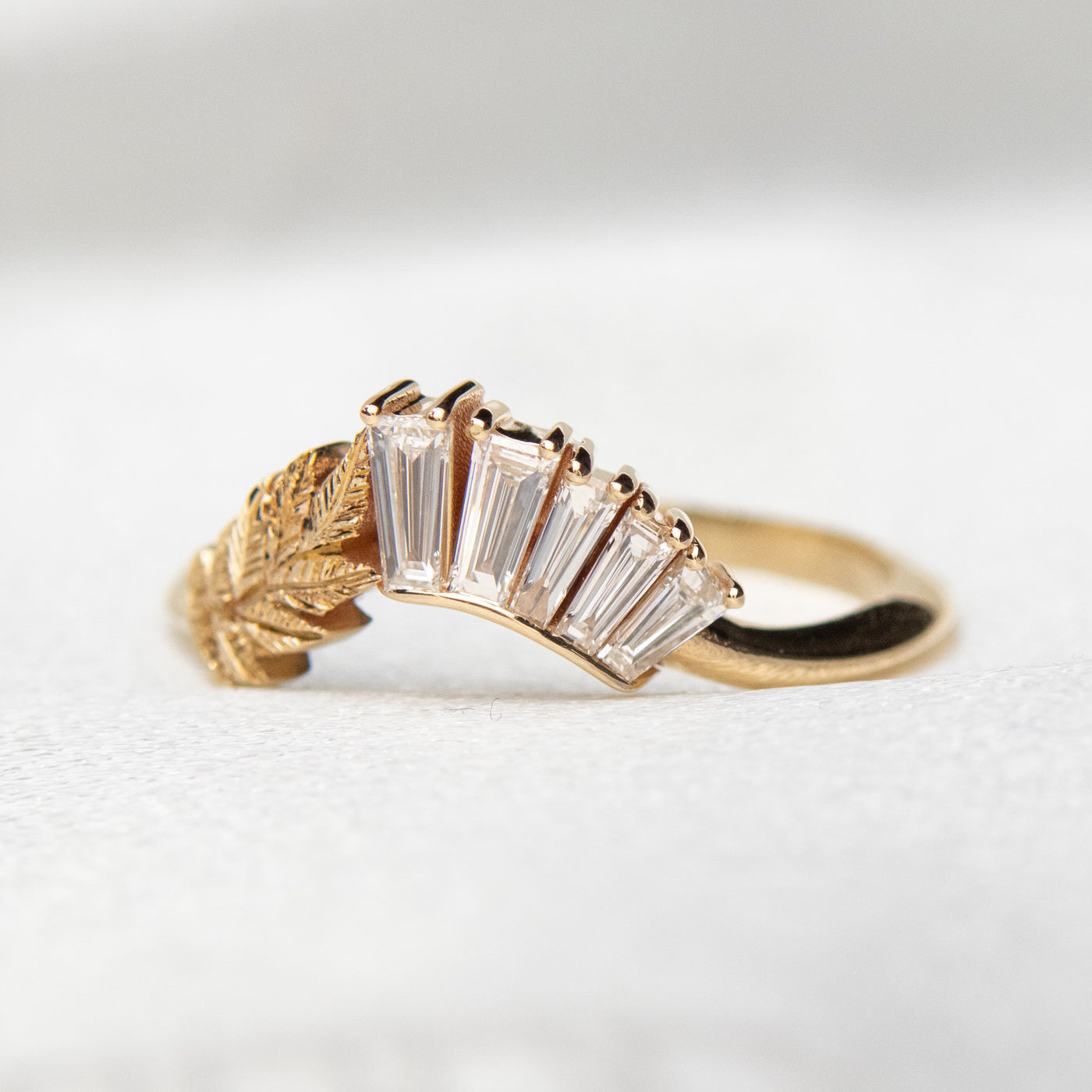 A slightly curved gold ring featuring a hand engraved leaf design and a row of sparkling, baguette cut lab grown diamonds - it can be worn as an wedding band or stacked with other rings.