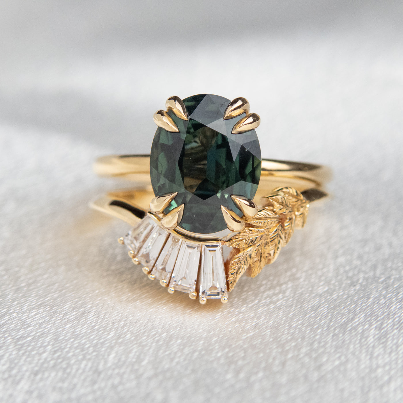 A slightly curved 14kt gold ring featuring a hand engraved leaf design and a row of sparkling, baguette cut lab grown diamonds stacked with a large green sapphire solitaire engagement ring in 14kt gold.