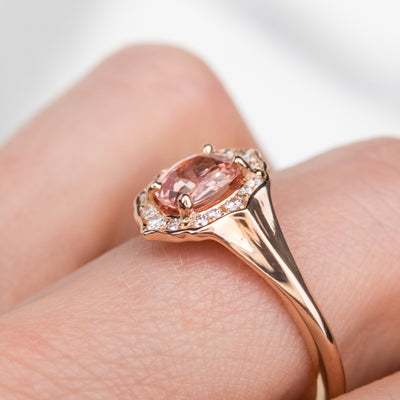 Elegant ring with an oval brilliant cut pink sapphire surrounded by a halo of small diamonds set in rose gold, worn on a hand.