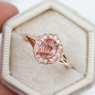 Elegant ring with an oval brilliant cut pink sapphire surrounded by a halo of small diamonds set in rose gold, against a ligt velvet ring box. 