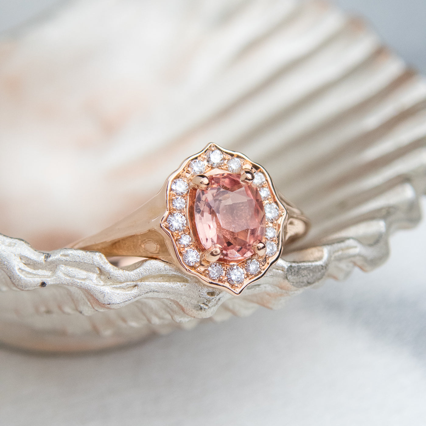 Elegant ring with an oval brilliant cut pink sapphire surrounded by a halo of small diamonds set in rose gold, resting on a silver seashell. 