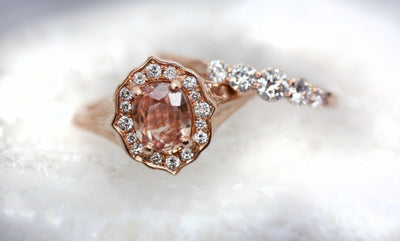 Elegant ring with an oval brilliant cut pink sapphire surrounded by a halo of small diamonds set in rose gold, against a white background. 