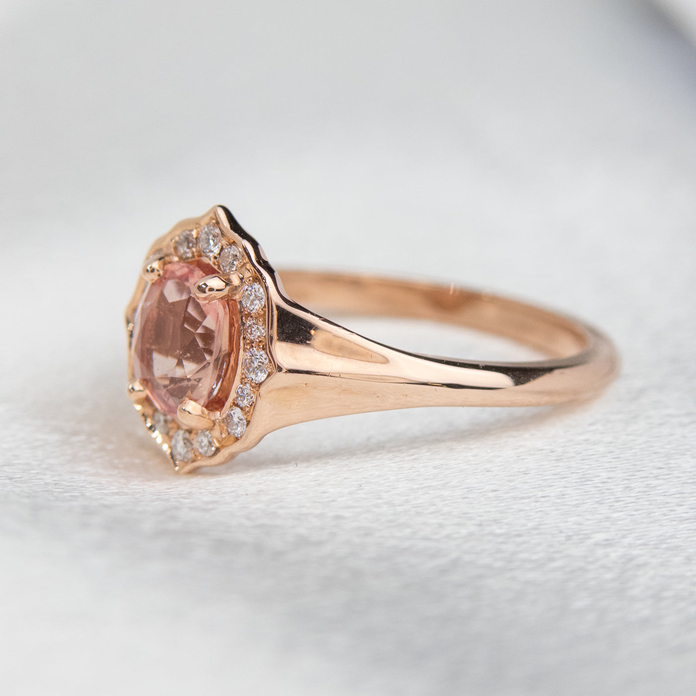 Elegant ring with an oval brilliant cut pink sapphire surrounded by a halo of small diamonds set in rose gold, against a soft, blurred background. 