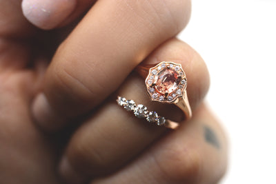 Elegant ring with an oval brilliant cut pink sapphire surrounded by a halo of small diamonds set in rose gold, paired with a graduated diamond band. Modelled on a hand.