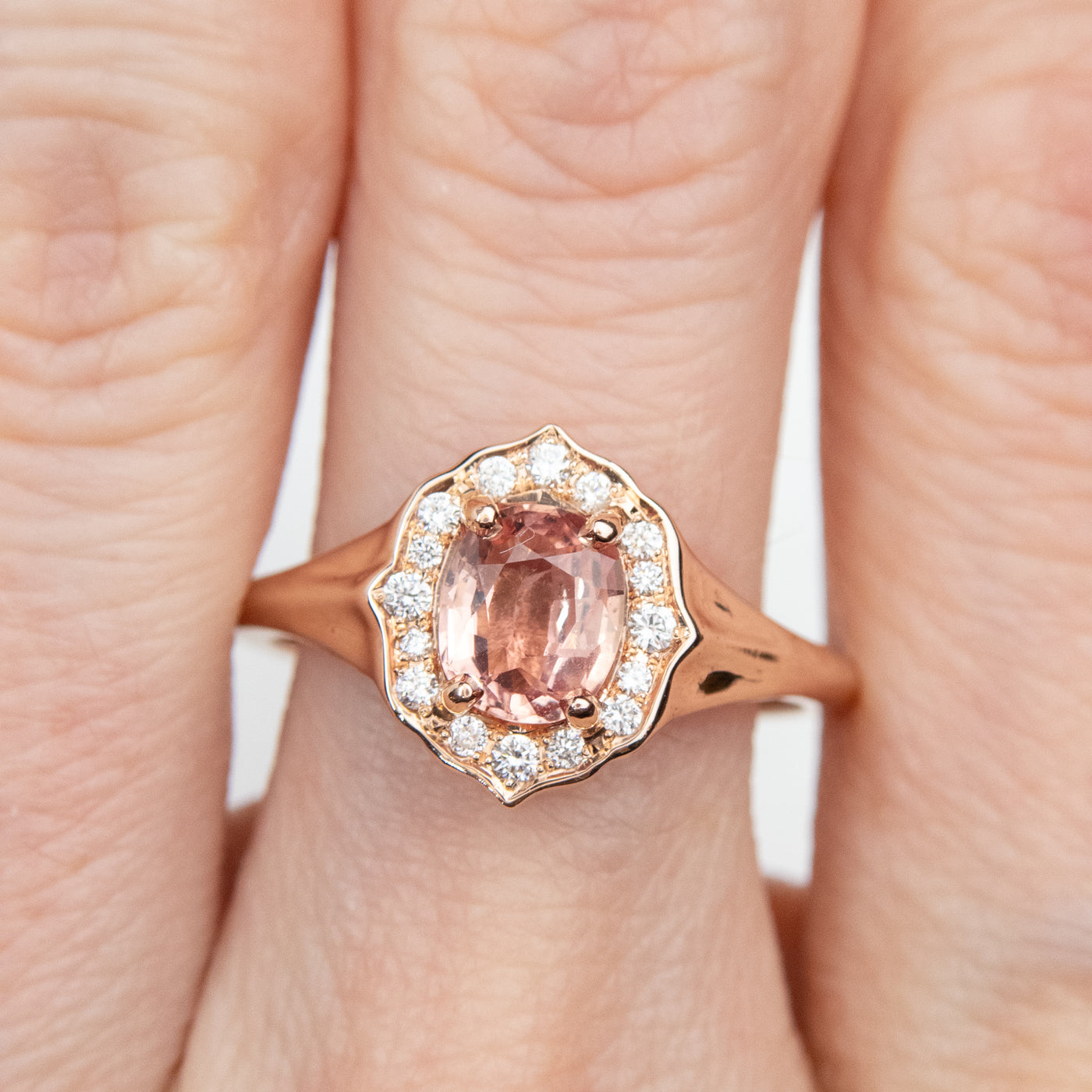 Elegant ring with an oval brilliant cut pink sapphire surrounded by a halo of small diamonds set in rose gold, worn on a hand.