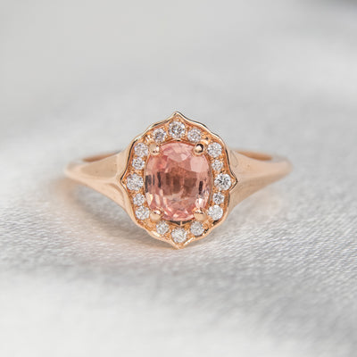 Elegant ring with an oval brilliant cut pink sapphire surrounded by a halo of small diamonds set in rose gold, against a soft, blurred background. 