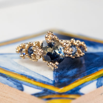 A gold engagement ring featuring a large, faceted blue sapphire center stone surrounded by intricate gold detailing and smaller sapphires, set on a colorful blue and yellow surface.