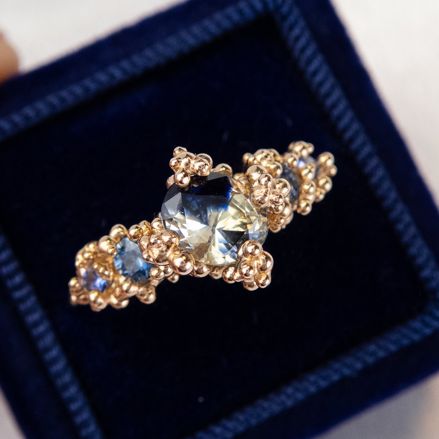 A gold engagement ring featuring a large, faceted blue sapphire center stone surrounded by intricate gold detailing and smaller sapphires, in a dark blue ring box. 