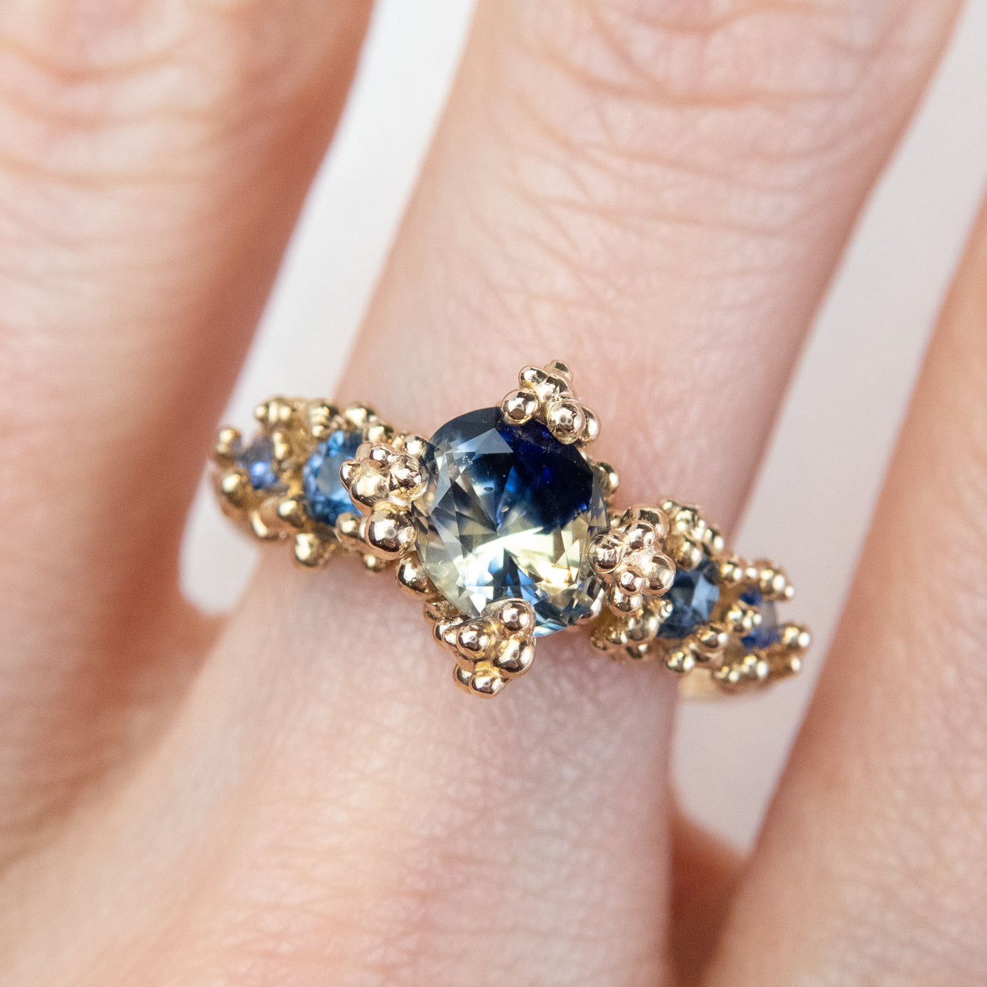 gold engagement ring featuring a large, faceted blue and yellow sapphire center stone surrounded by intricate gold detailing and smaller sapphires, shown on a finger.