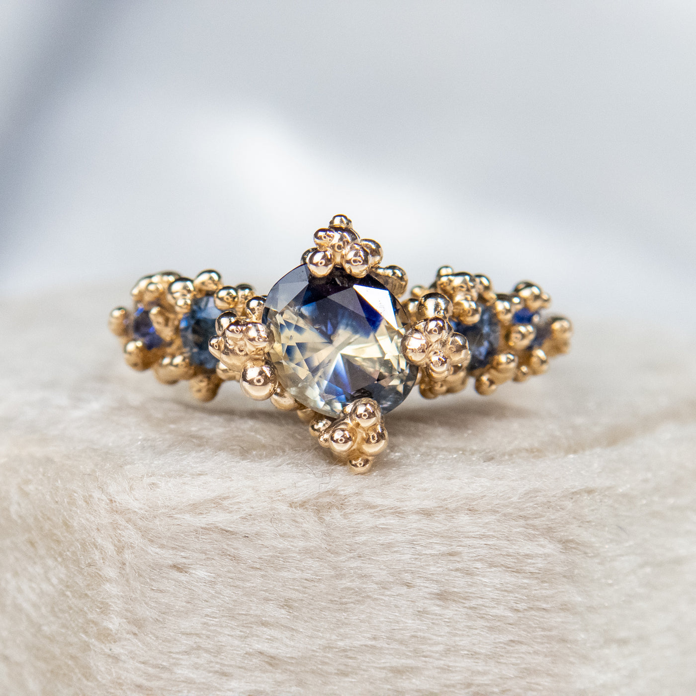 Gold engagement ring featuring a large, faceted blue sapphire center stone surrounded by intricate gold detailing and smaller sapphires, set on a plush, light-colored surface.