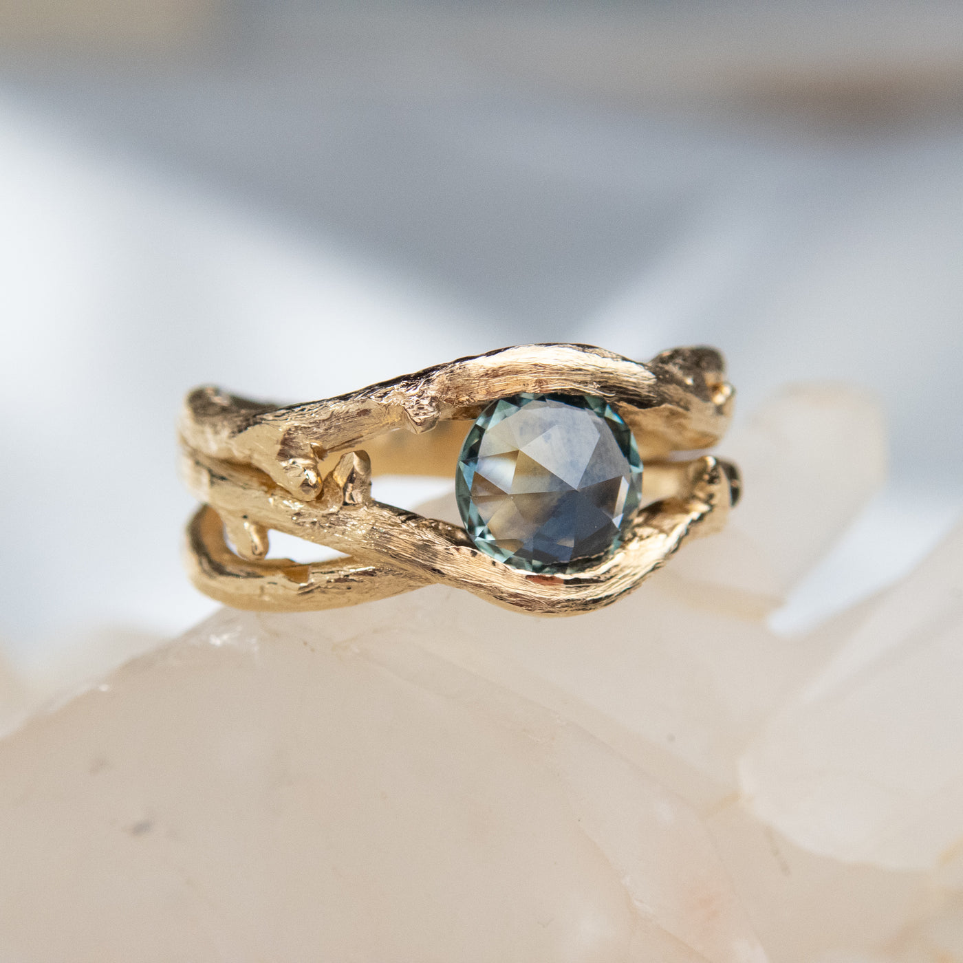 Gold ring with a unique twisted band design, featuring a round rose cut teal blue sapphire, displayed on a white crystal background.