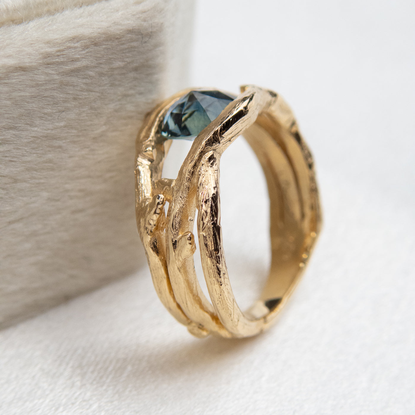 Side view of a gold ring with a unique twisted band design, featuring a round rose cut teal blue sapphire, displayed on a light fabric background.