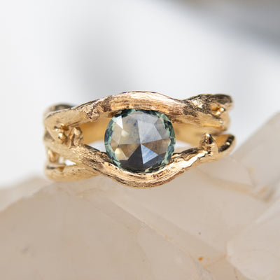 Gold ring with a unique twisted band design, featuring a round rose cut teal blue sapphire, displayed on a white crystal background.