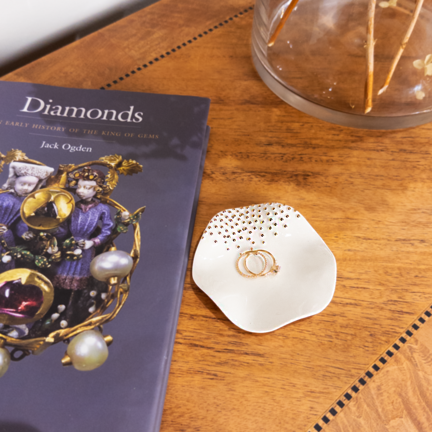 Two elegant gold rings are displayed on a smooth, white, dish-shaped jewelry tray adorned with small golden beads resting on a table next to a book to show scale.