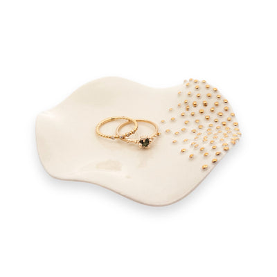 Two elegant gold rings are displayed on a smooth, white, dish-shaped jewelry tray adorned with small golden beads.