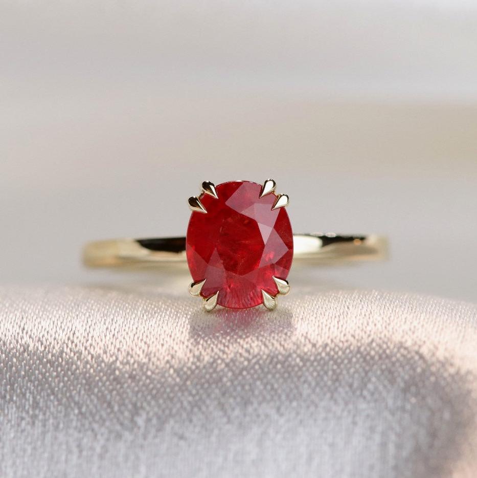 A close-up view of a gold ring featuring an oval-cut red sapphire, elegantly set in delicate double claw prongs. 
