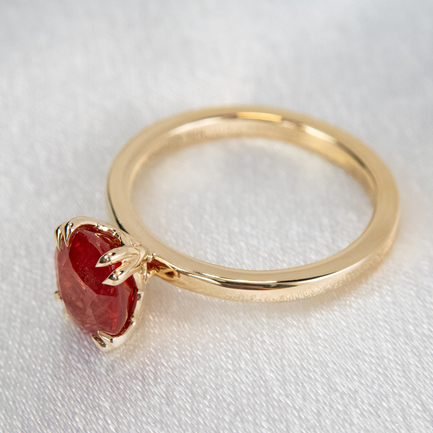 A gold ring featuring an oval-cut red sapphire, elegantly set in delicate double claw prongs. It rests on a soft, light fabric surface.