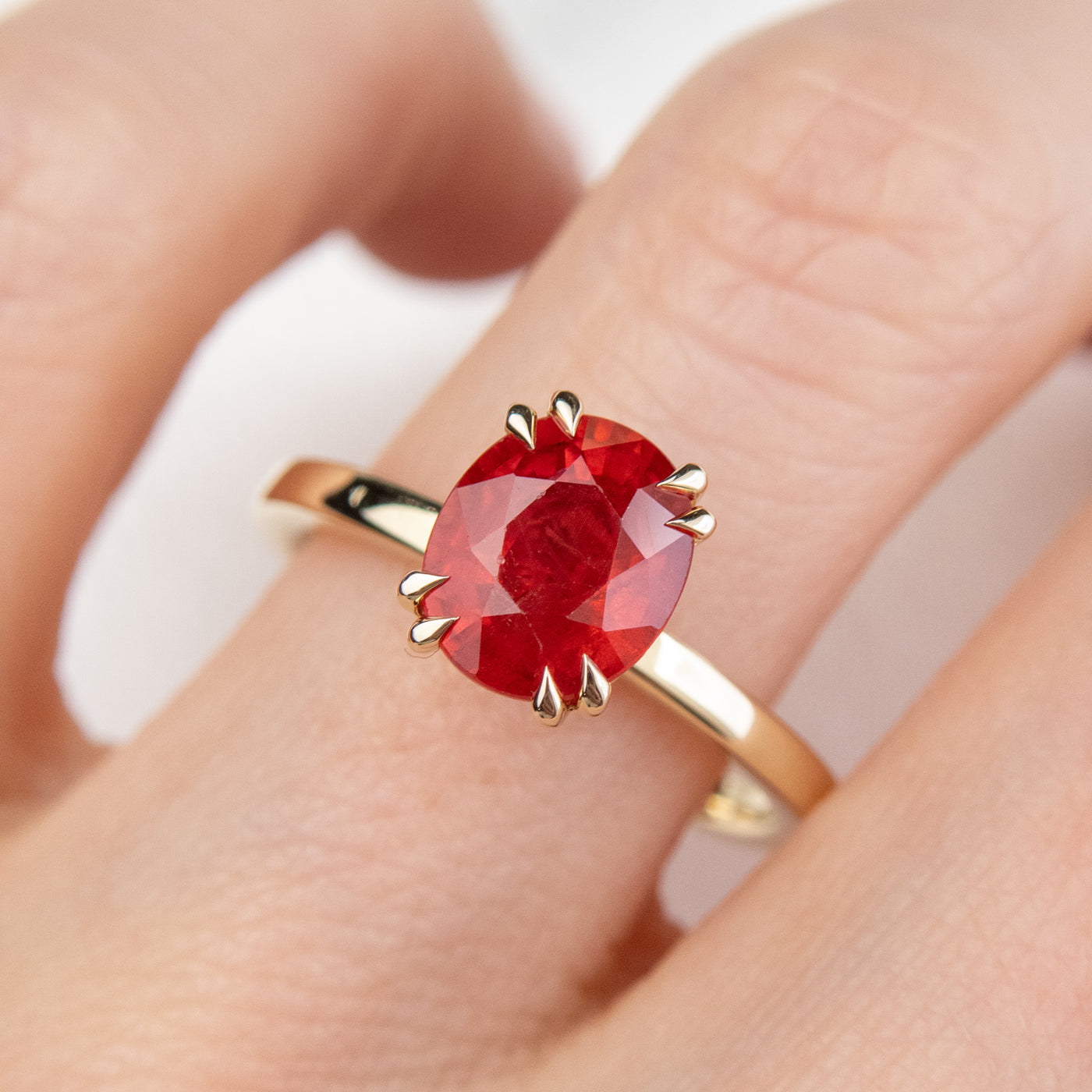 A gold ring featuring an oval-cut red sapphire, elegantly set in delicate double claw prongs. Worn on a finger. 