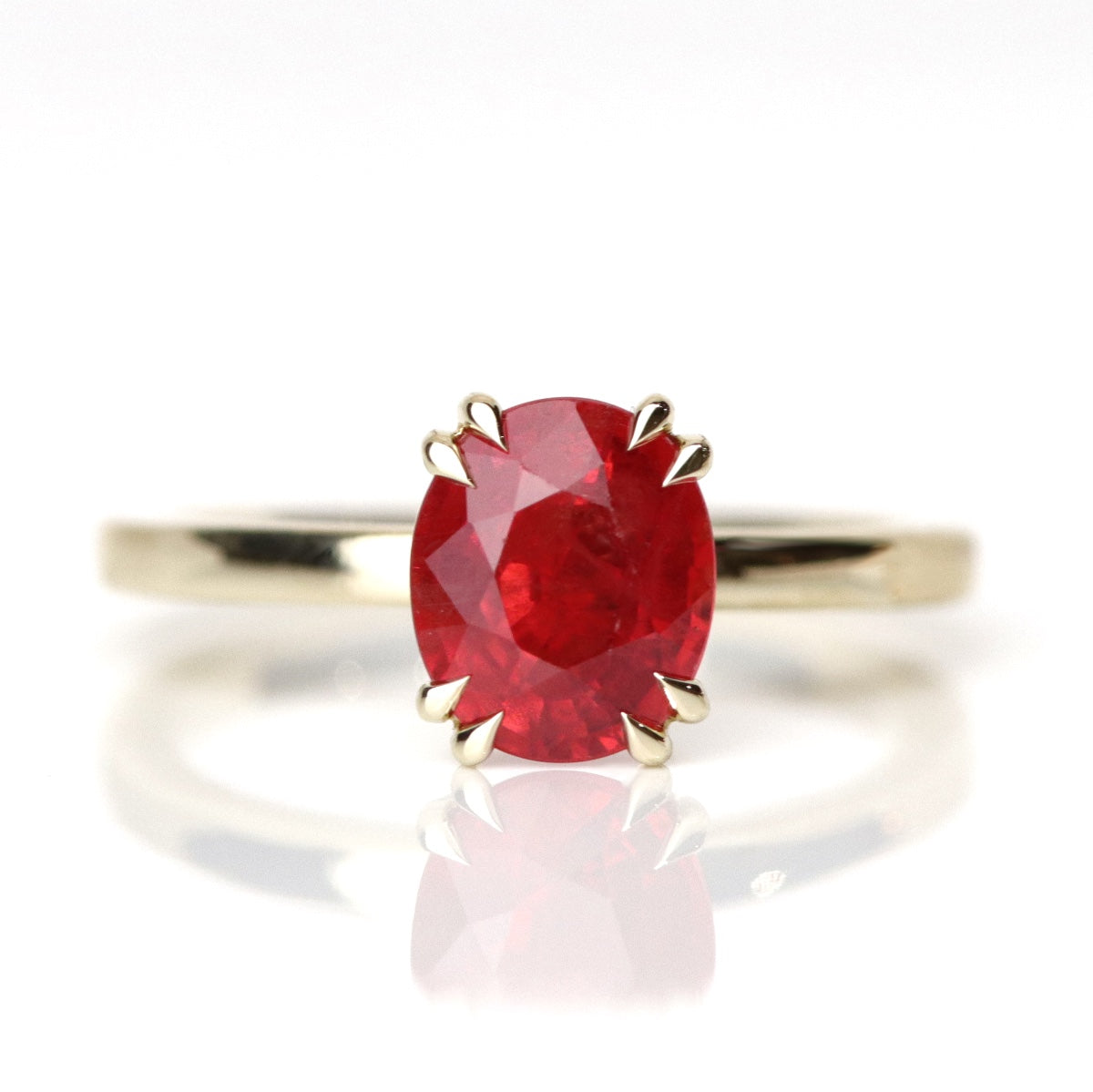 A gold ring featuring an oval-cut red sapphire, elegantly set in delicate double claw prongs. Pictured against a reflective white background. 