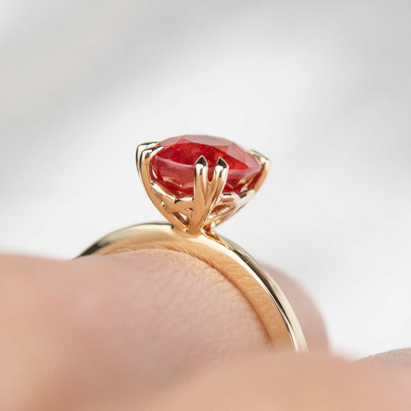 A close-up side view of a gold ring featuring an oval-cut red sapphire, elegantly set in delicate double claw prongs. Worn on a finger. 