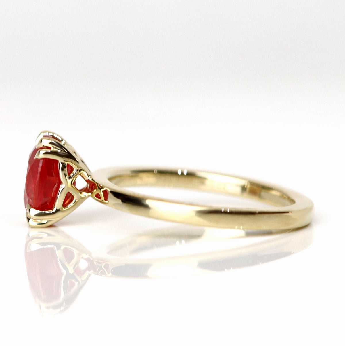 A side view of a gold ring featuring an oval-cut red sapphire, elegantly set in delicate double claw prongs. Pictured on a reflective white background. 