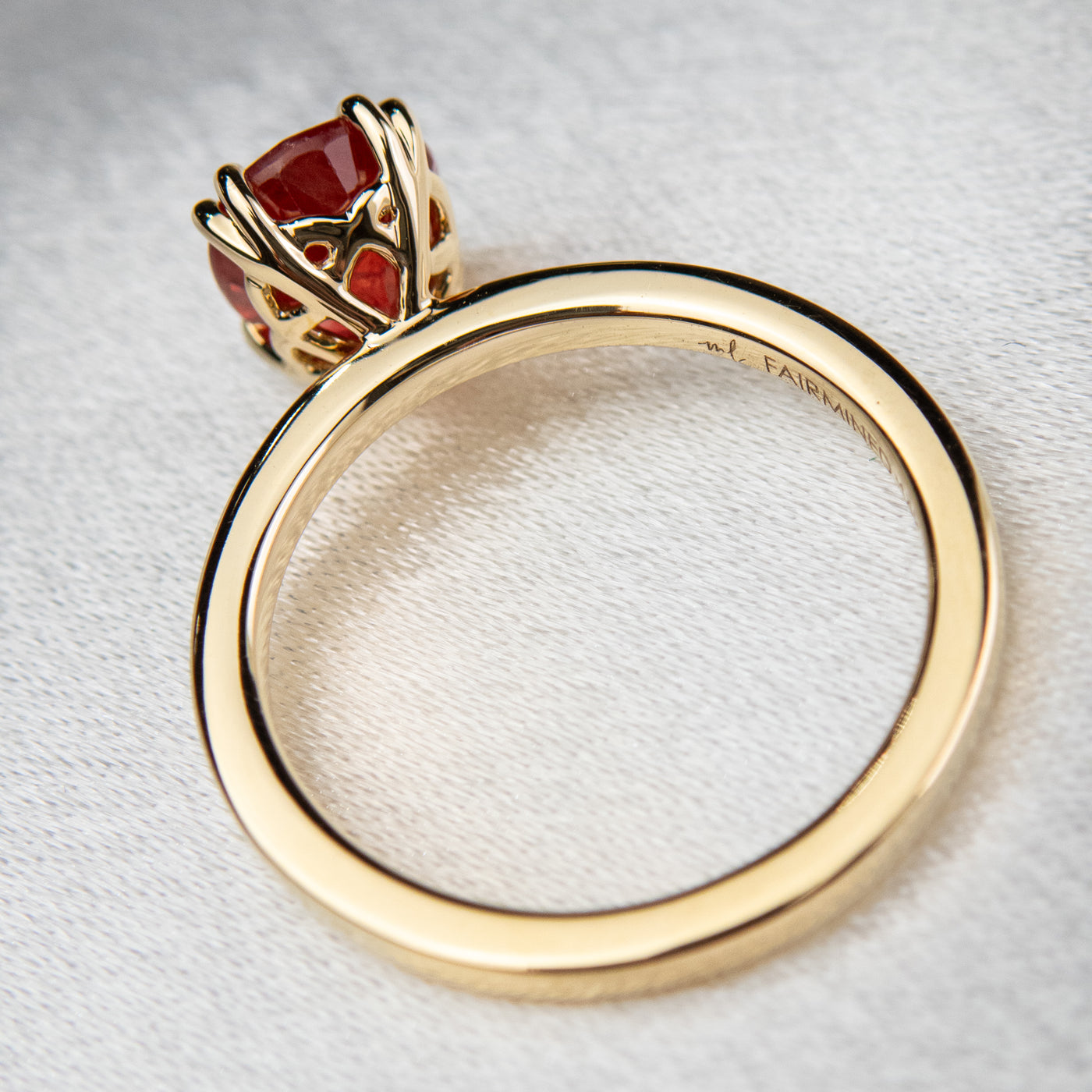 A side view of a gold ring featuring an oval-cut red sapphire, elegantly set in delicate double claw prongs with an intricate basket setting. It rests on a soft, light fabric surface.