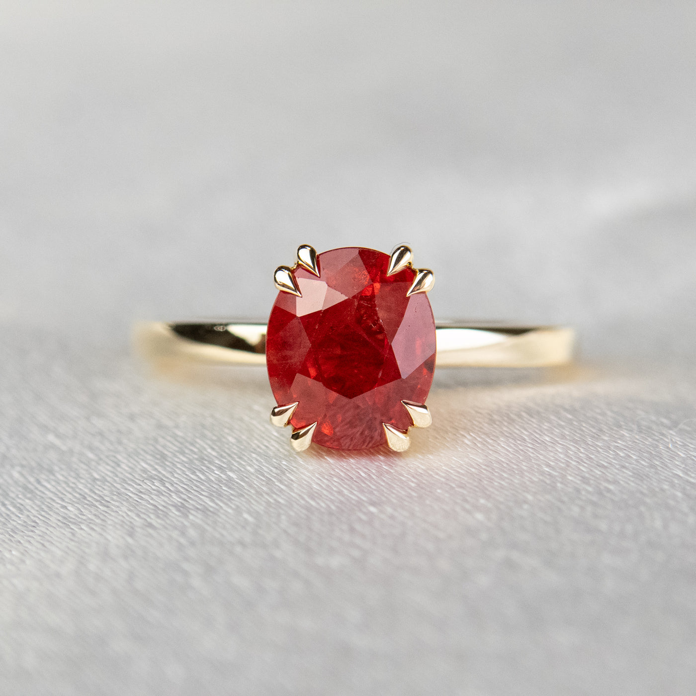 A gold ring featuring an oval-cut red sapphire, elegantly set in delicate double claw prongs. It rests on a soft, light fabric surface.