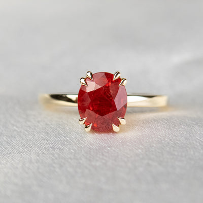 A gold ring featuring an oval-cut red sapphire, elegantly set in delicate double claw prongs. It rests on a soft, light fabric surface.