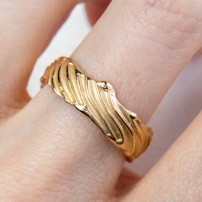 A 14kt gold wedding ring with intricate wave-like engravings, shown worn elegantly on a finger.