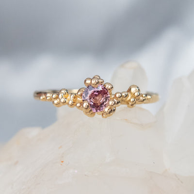 A gold ring with a cluster of small round gold beads holds a pink sapphire at the center, displayed on a white crystalline surface.