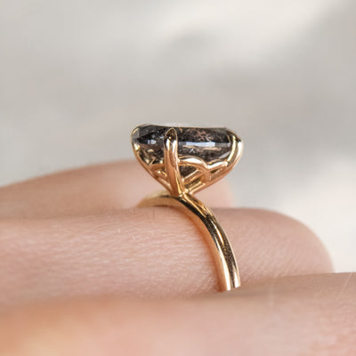 Close-up side view of an elegant gold ring set with a large oval salt and pepper diamond. It features tiger-style prongs atop a decorative under basket, worn on a hand. 